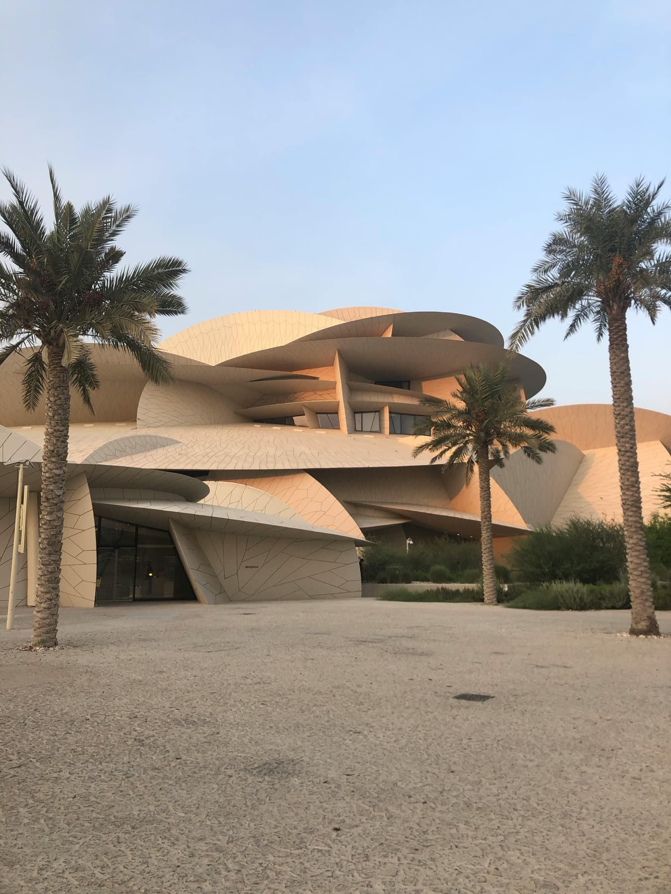 Sand-toned contemporary building with interlocking curved discs, framed by date palms under a soft sky in Doha.